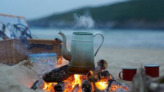 An old kettle boils on a small bonfire at the beach with a picnic basket in the background.