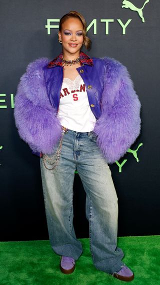 Rihanna wearing a purple fur coat with a varsity-style tee and low-rise jeans with a bejeweled chain