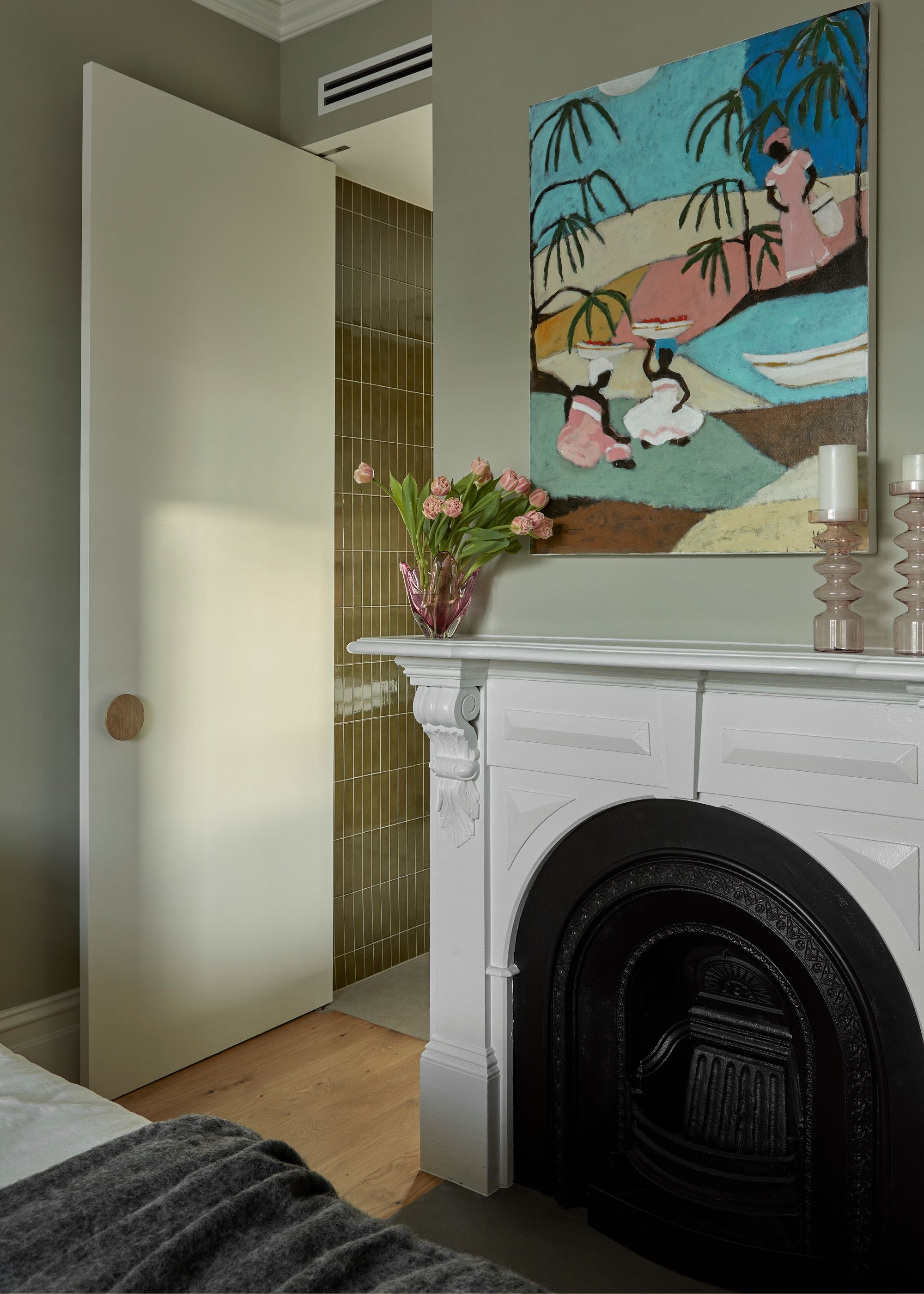 A bedroom with a white and black fireplace, a vase of tulips, a colorful painting, and a pair of candles on holders