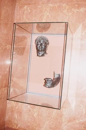 Bronze statue in two parts - head and left arm - in a glass box displayed against a terracotta marble background.