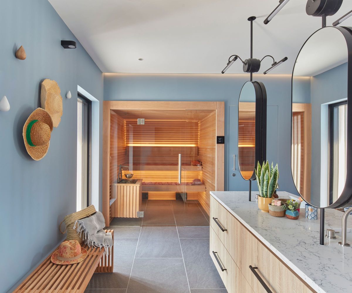 A blue-painted pool house changing room with a glowing cedar sauna visible through a glass door, a marble-topped oak vanity, oval mirror and straw hats on the wall.