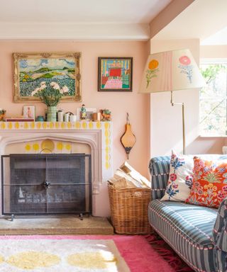 colorful living room with pink and yellow rug, blue patterned couch, cushions, pink upholstered chair, artwork, fireplace that's painted, painted lampshade on floor lamp