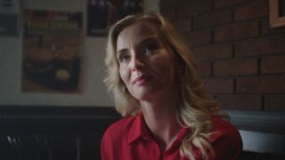 Amber sitting in a dark dive bar with orange brickwork in the background, dressed in a bright red blouse looking longingly at Flynn, who is off screen.