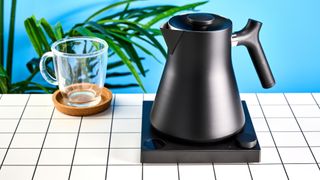 the fellow Corvo EKG kettle in matte black photographed against a blue background with a glass mug and black and white table 