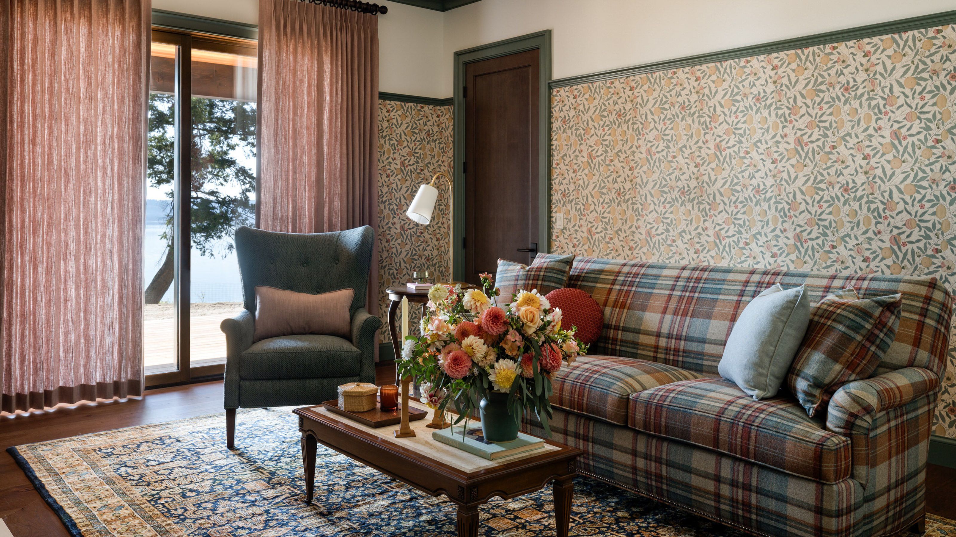 A snug room with muted floral wallpaper, green trim, warm white walls, muted pink curtains on a glass sliding door, a patterned rug, and a tartan sofa.