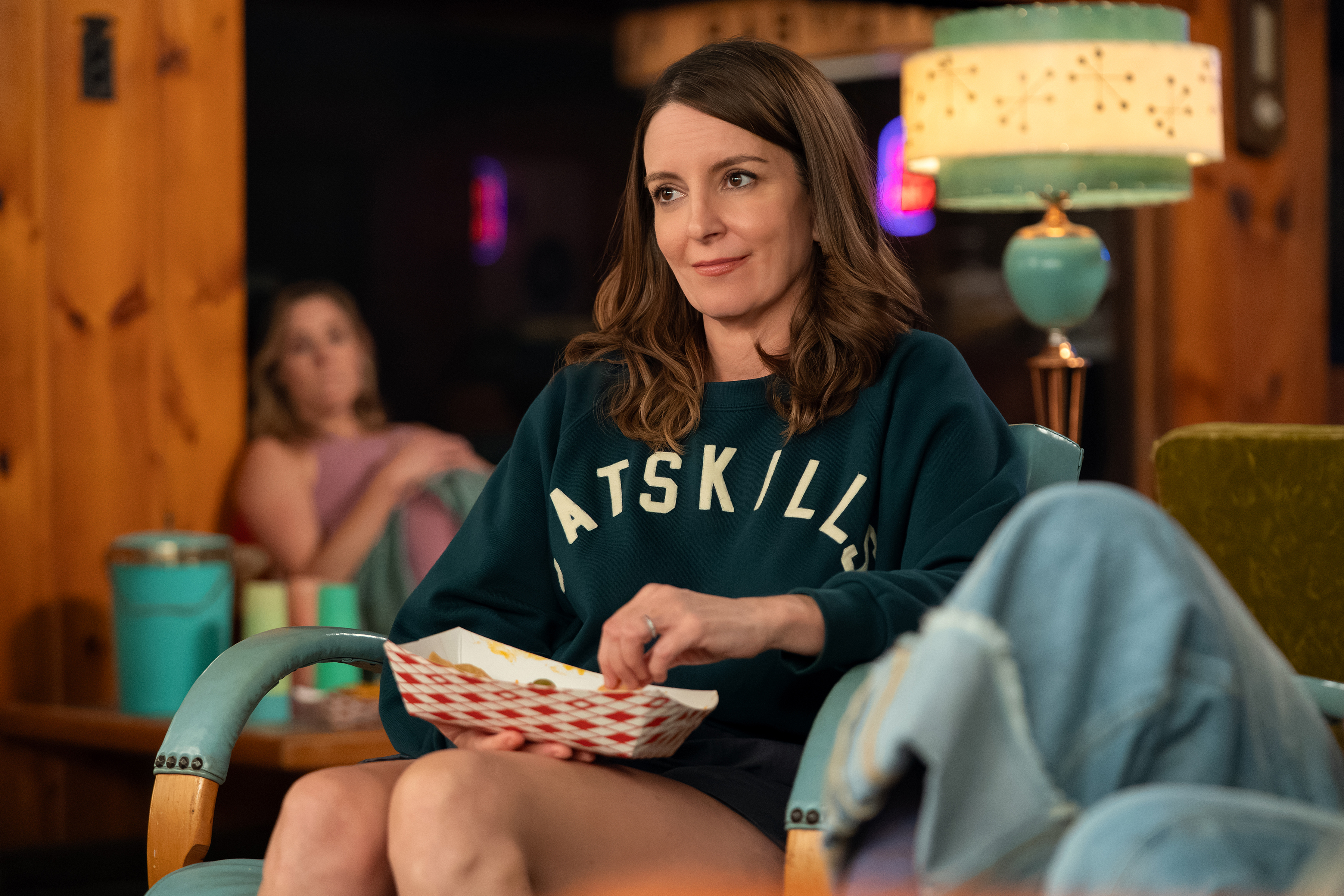 Tina Fey as Kate wearing a catskills sweatshirt eating a snack in a still from the four season season 2