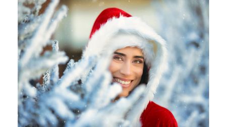 A model dressed as Santa Claus smiles from behind a frosted pine tree