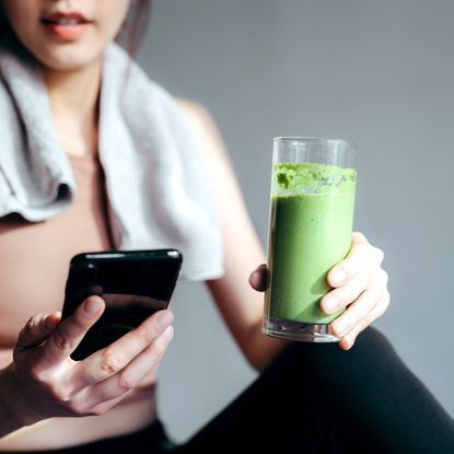 close up of confidence young asian sports woman taking a break, refreshing with healthy green juice and using smartphone after fitness work out exercising practicing yoga at home in the fresh bright morning