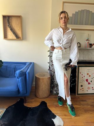 woman standing in front of apartment wearing white button down, white denim mid skirt with slit, white socks and green Adidas sneakers, pearl necklace