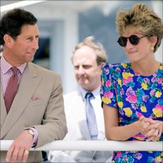 King Charles looks at Princess Diana who is wearing a blue floral dress featuring pink, yellow, and purple flowers with round sunglasses