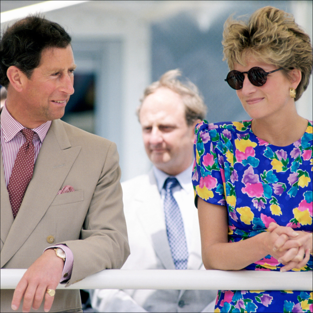 King Charles looks at Princess Diana who is wearing a blue floral dress featuring pink, yellow, and purple flowers with round sunglasses