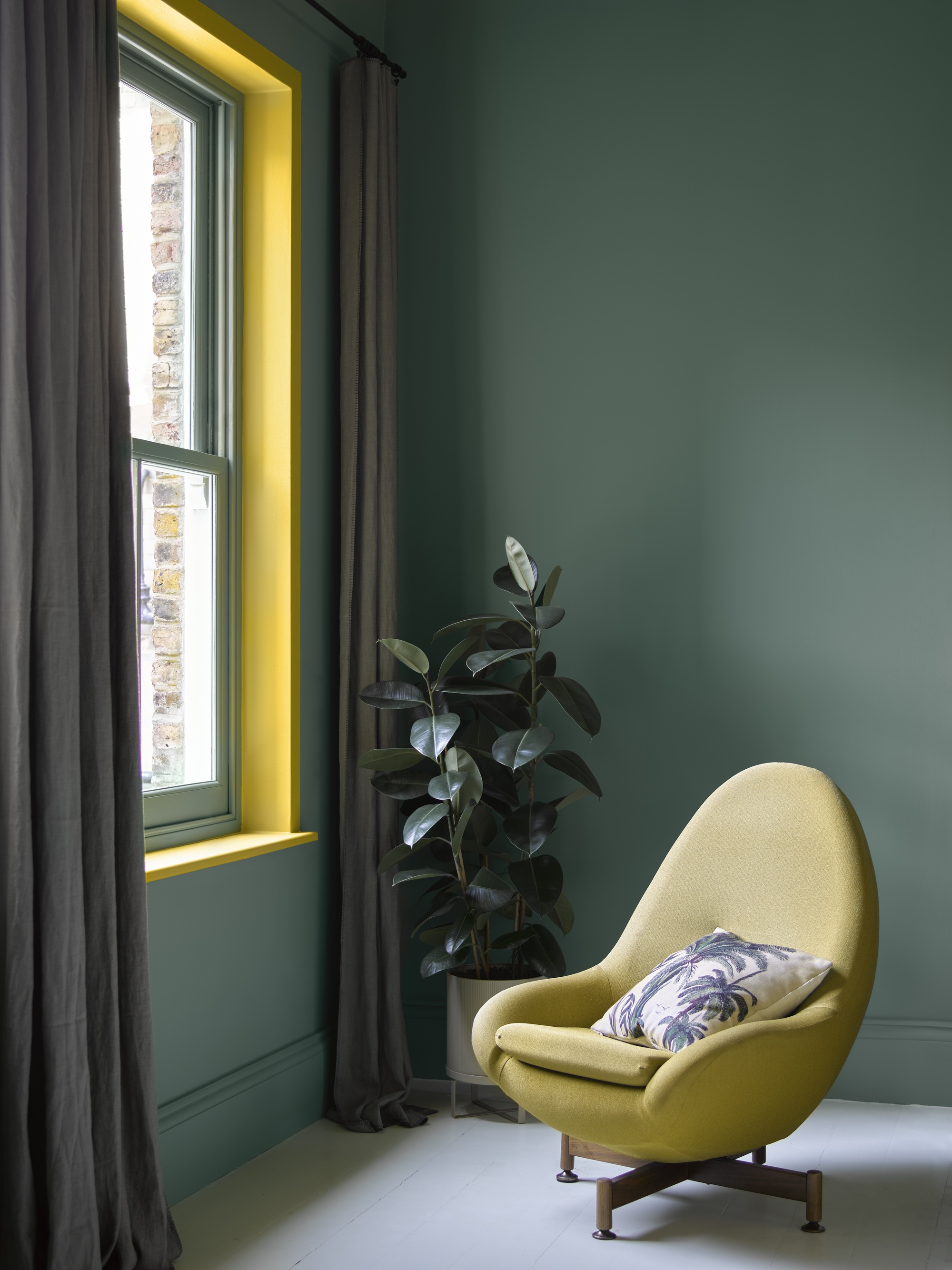 Image of the corner of a room painted in a dark teal with a window that has a bright yellow frame and gray curtains. There is a plant and a yellow egg chair.