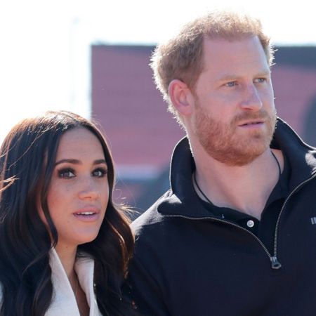 Meghan Markle has long brown hair and is wearing a white jacket while she stands with husband Prince Harry, who wears a black polo shirt with the Invictus Games logo