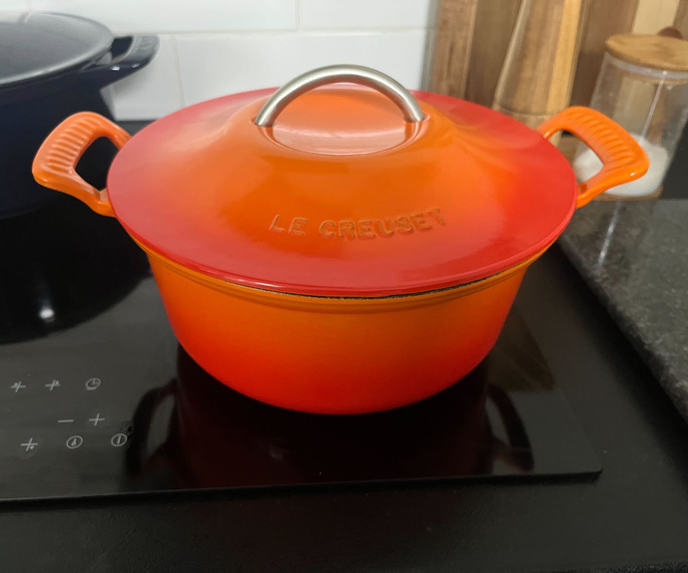 An orange cast iron Dutch oven with flared side handles, and a lid with a curved metal handle on top. On a black electric stovetop
