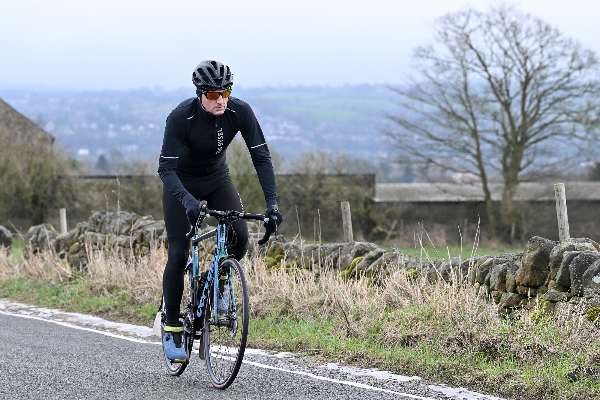 Front 3/4 of a man riding out of the saddle wearing a black jacket, black tights and a black helmet on a green Cervelo bike