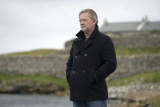 Douglas Henshall as DI Jimmy Perez in Shetland, standing on the seafront and staring out to sea, looking troubled. He is wearing his trademark black double-breasted peacoat, and has his hands in his jacket pockets. Behind him, out of focus, there are dry stone walls and a cottage