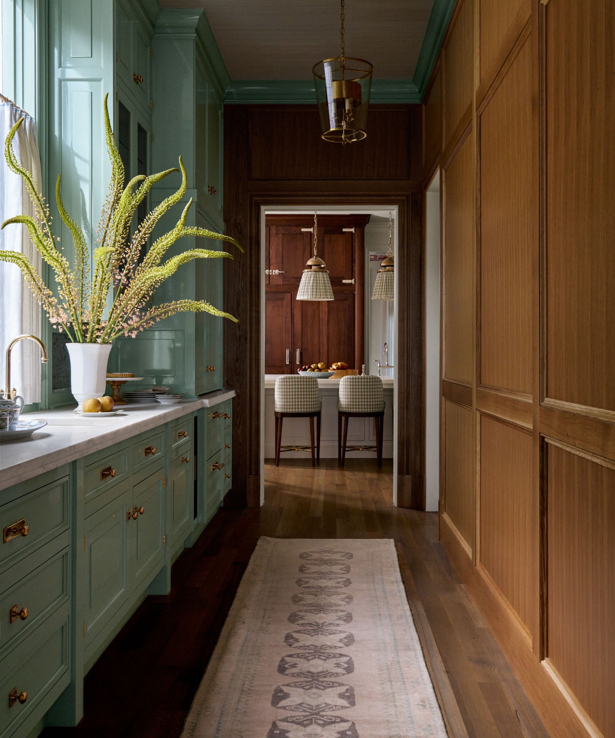 A pass-through galley kitchen with mint green lacquered cabinetry, a marble countertop with a sink and a large vase of foliage; on the left is dark brown cabinetry leading down into a larger kitchen.