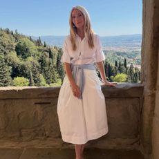 Gwyneth Paltrow poses for a photo outside wearing a white cotton button-down dress with a sky blue jumper wrapped around her waist and a pair of sky blue heels.