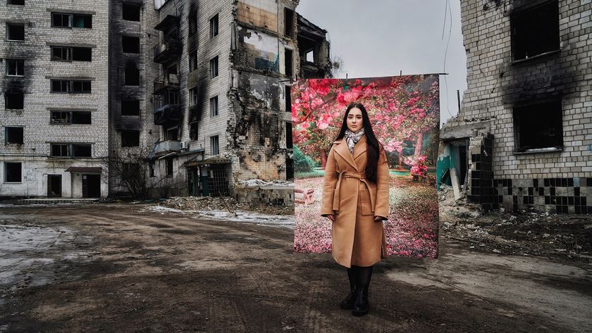 A woman in a tan coat stands in front of a backdrop depicting a blooming spring scene, flanked by heavily damaged, multi-story buildings.