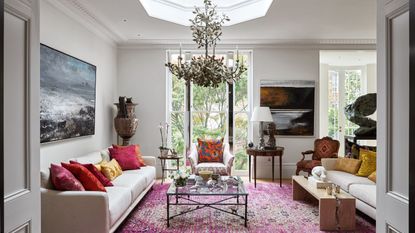 A white living room with large pink rug, white sofa with red and yellow cushions, a statement lamp and wooden furniture. On the walls are large painted seascapes.