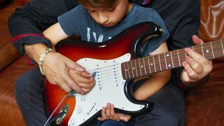 Young boy sits on his father's lap while playing a full-size guitar