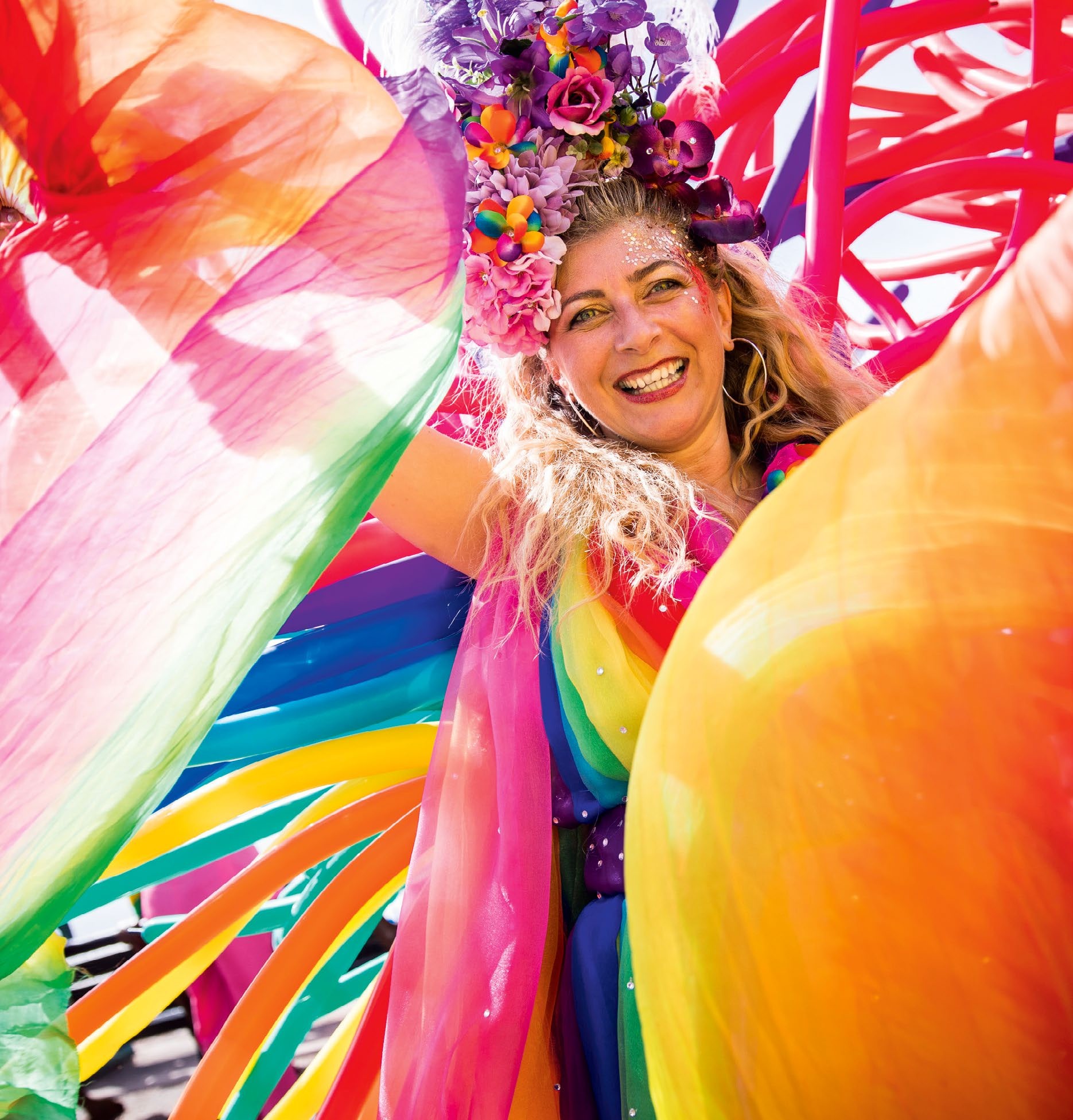 A joyful woman in vibrant rainbow attire and a floral headdress, celebrating at a festival with colorful flowing ribbons.