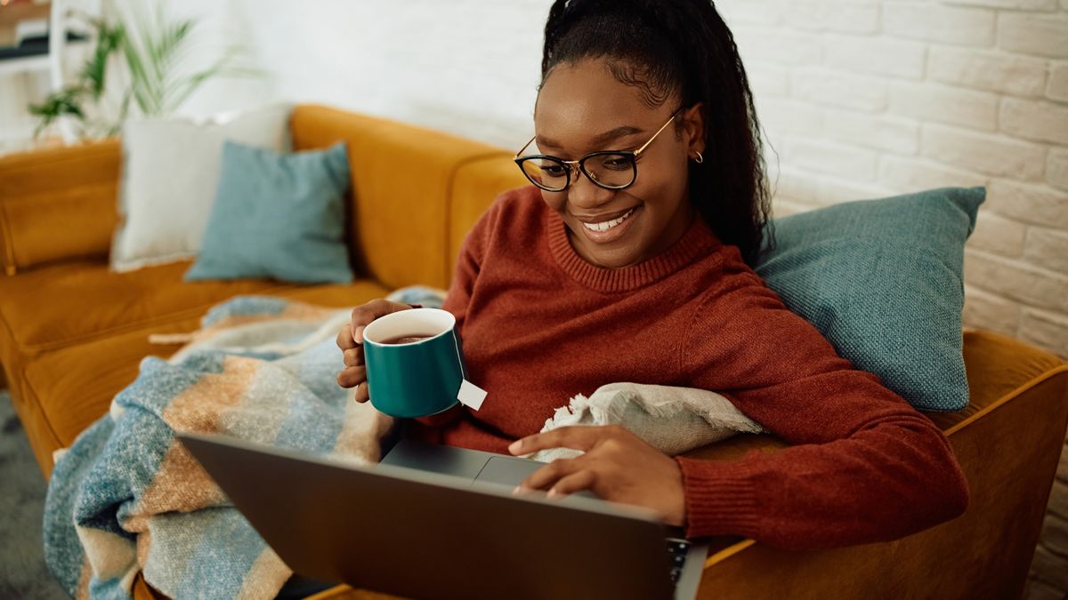 A woman sitting on a sofa with a throw, holding a cup of tea and using a laptop