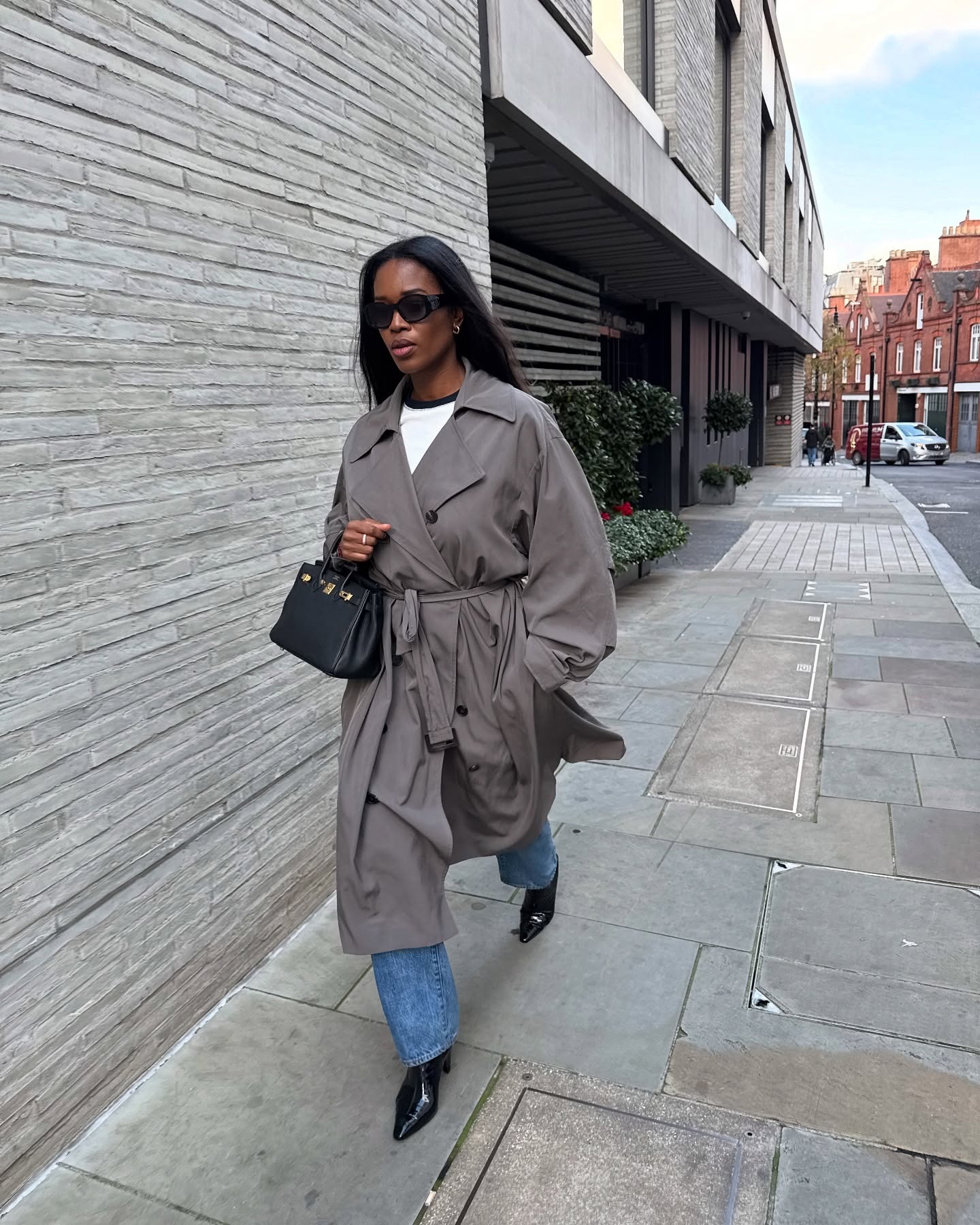 Influencer @nlmarilyn walks down the street in London wearing chisel-toe boots with jeans and a grey trench coat.