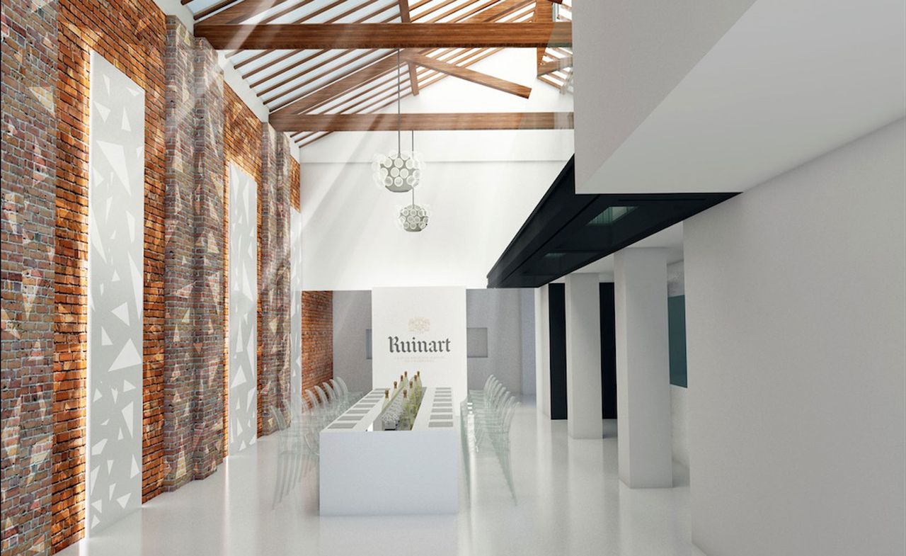 Interior view at The Ruinart Lighthouse show featuring white floors, white and brick walls with a triangle pattern projected onto one side, a long white table with bottles of drinks on top, clear chairs, pendant lights and the wording &#039;Ruinart&#039; on the back wall