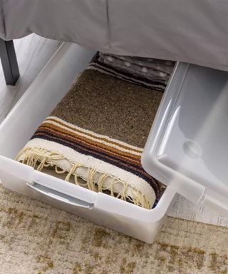 An image of a bed with a an open plastic storage crate with a rug in it underneath the bed