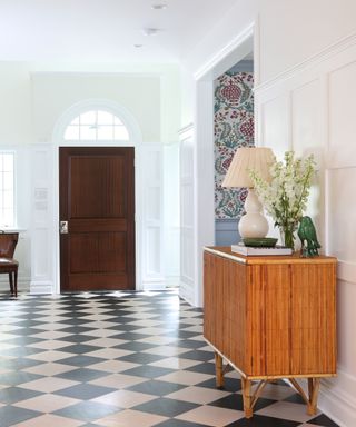 Large hallway with black and beige chequered stone flooring, white painted walls with wall paneling, bamboo style unit with table lamp and vase of flowers, dark wood door