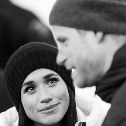 A black and white photo of Meghan Markle wearing a winter hat and coat looking up at Prince Harry