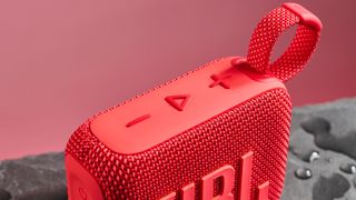 The play, pause, and volume buttons on the top of a red JBL Go 4 ,pictured against a pink background. It is sitting on a dark stone surface with puddles of water around it.