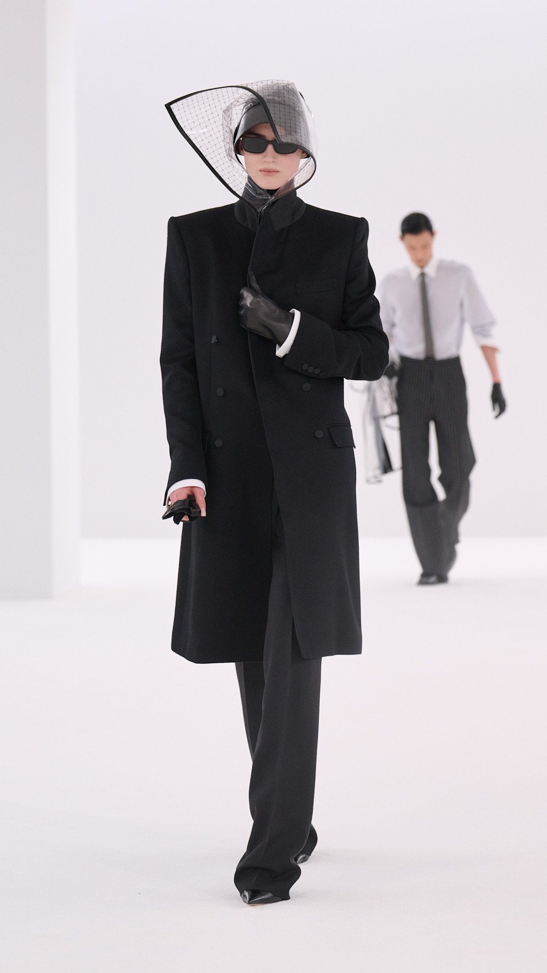 a Tom Ford model wearing a PVC cap, black coat, and black pants.