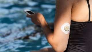 Woman sat on the side of a swimming pool with a continuous glucose monitor (CGM) in her arm, using her phone to check the real-time data