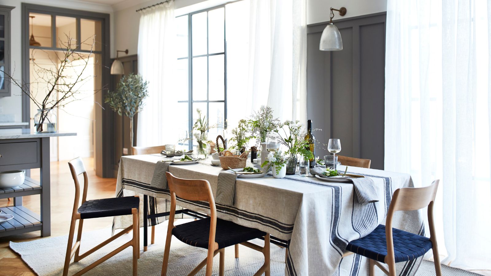 A bright and airy dining room with a striped tablecloth on a long table, set with wine glasses, wine, and napkins, and blue and brown chairs on top of a white rug. Behind are brown panelled walls, and sheer curtains over crittal windows.