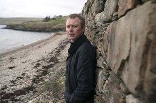 DC Sandy Wilson (Steven Robertson) stands on a pebbly beach, leaning against a brick wall behind him. His face is turned towards the camera.