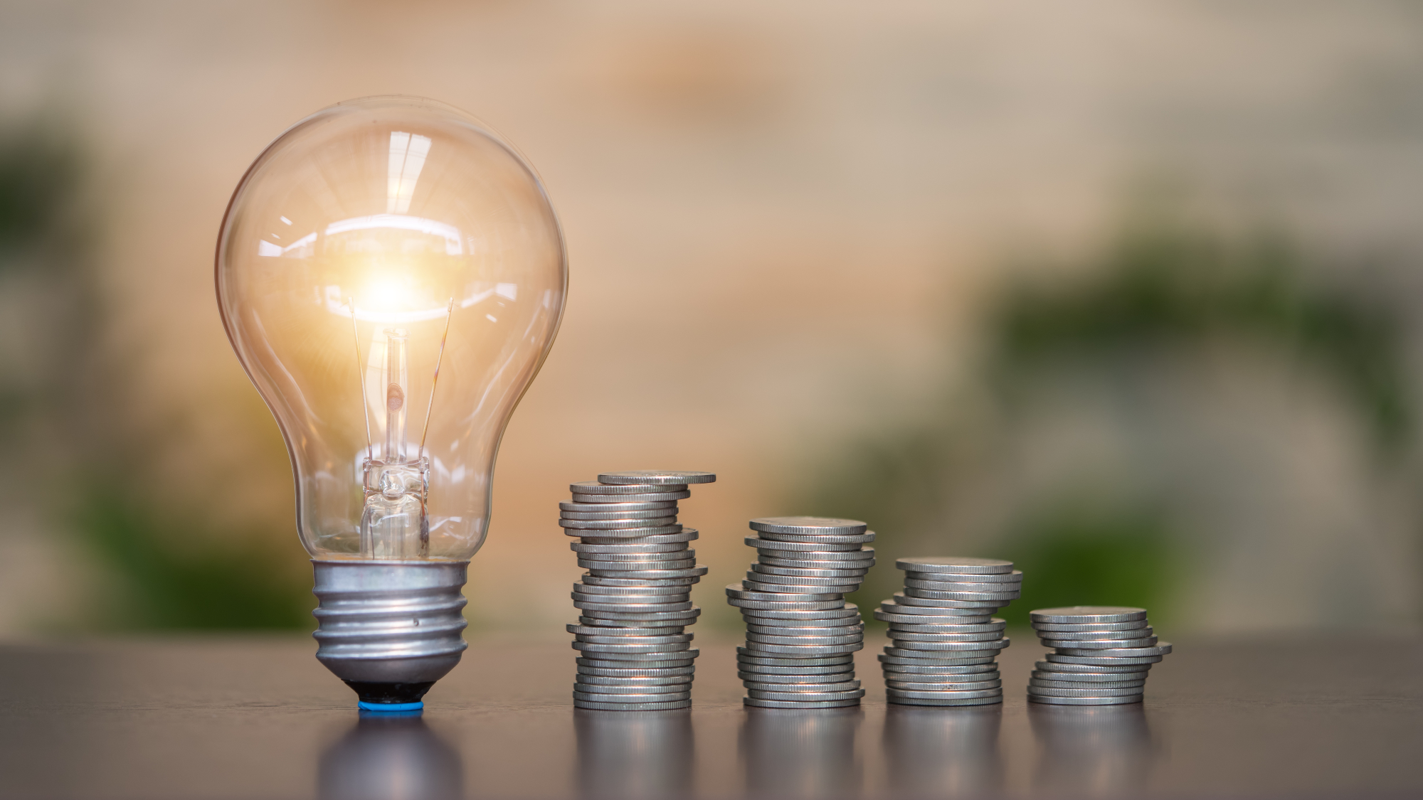 lit up light bulb on table with piles of coins next to it