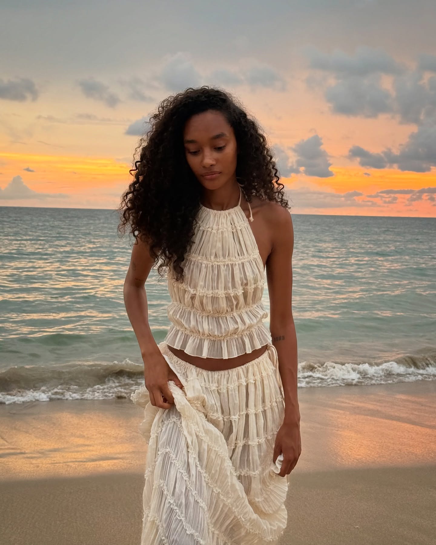 A woman walks on the beach wearing a two-piece white set with her hair down and naturally curly.