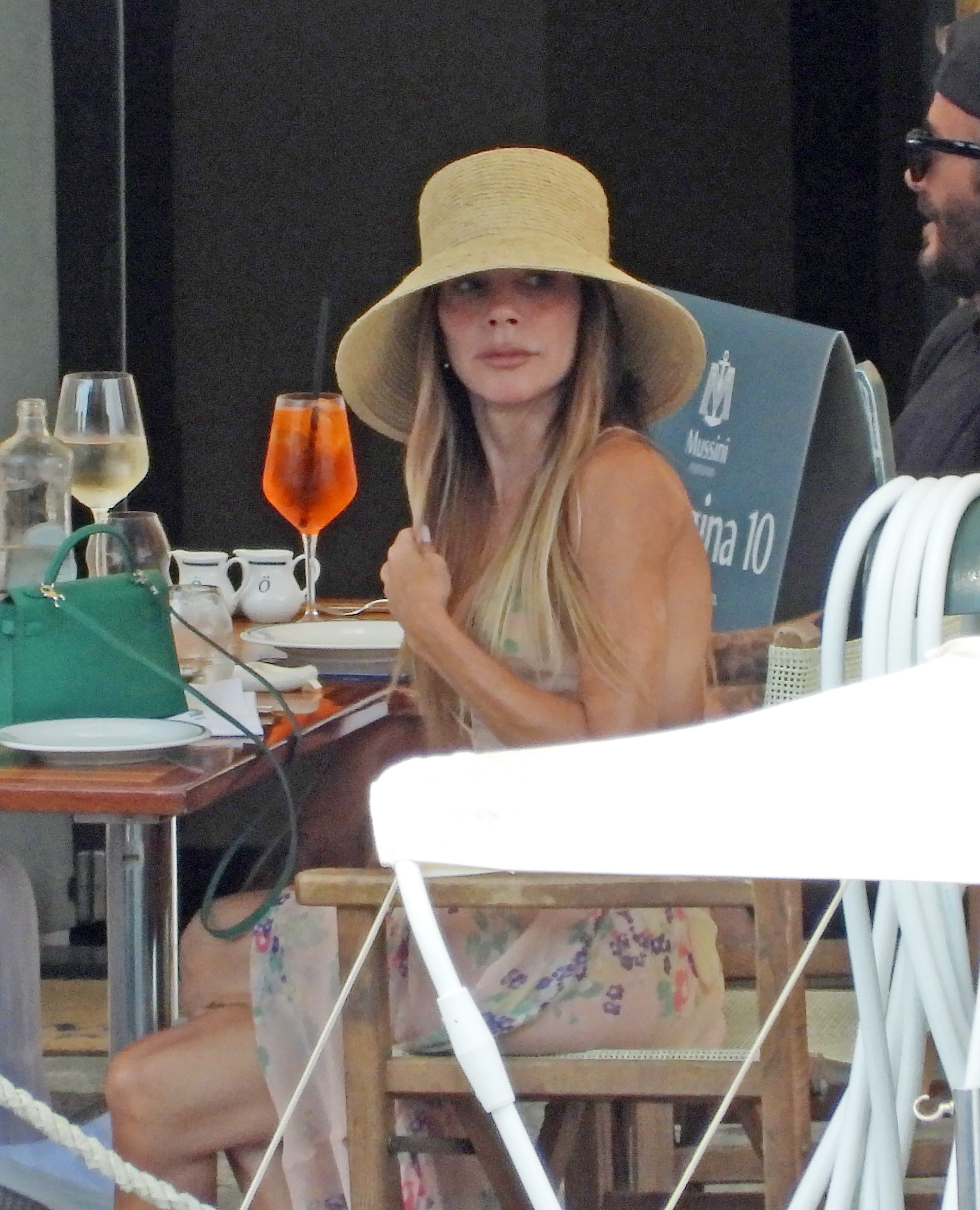 Victoria Beckham wears a sheer naked vacation dress paired with a massive hat and a green Hermes Kelly bag