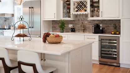 A white kitchen island, tiered stand with bread and cookies on it and a wooden fruit bowl, two white chairs underneath it, plus white cabinets with gold handles