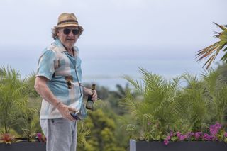 Gerry Stableforth (Geoff Bell) stands on his balcony surrounded by exotic plants, wearing a hat and sunglasses. He has a beer bottle in his left hand and his mobile phone in his right.