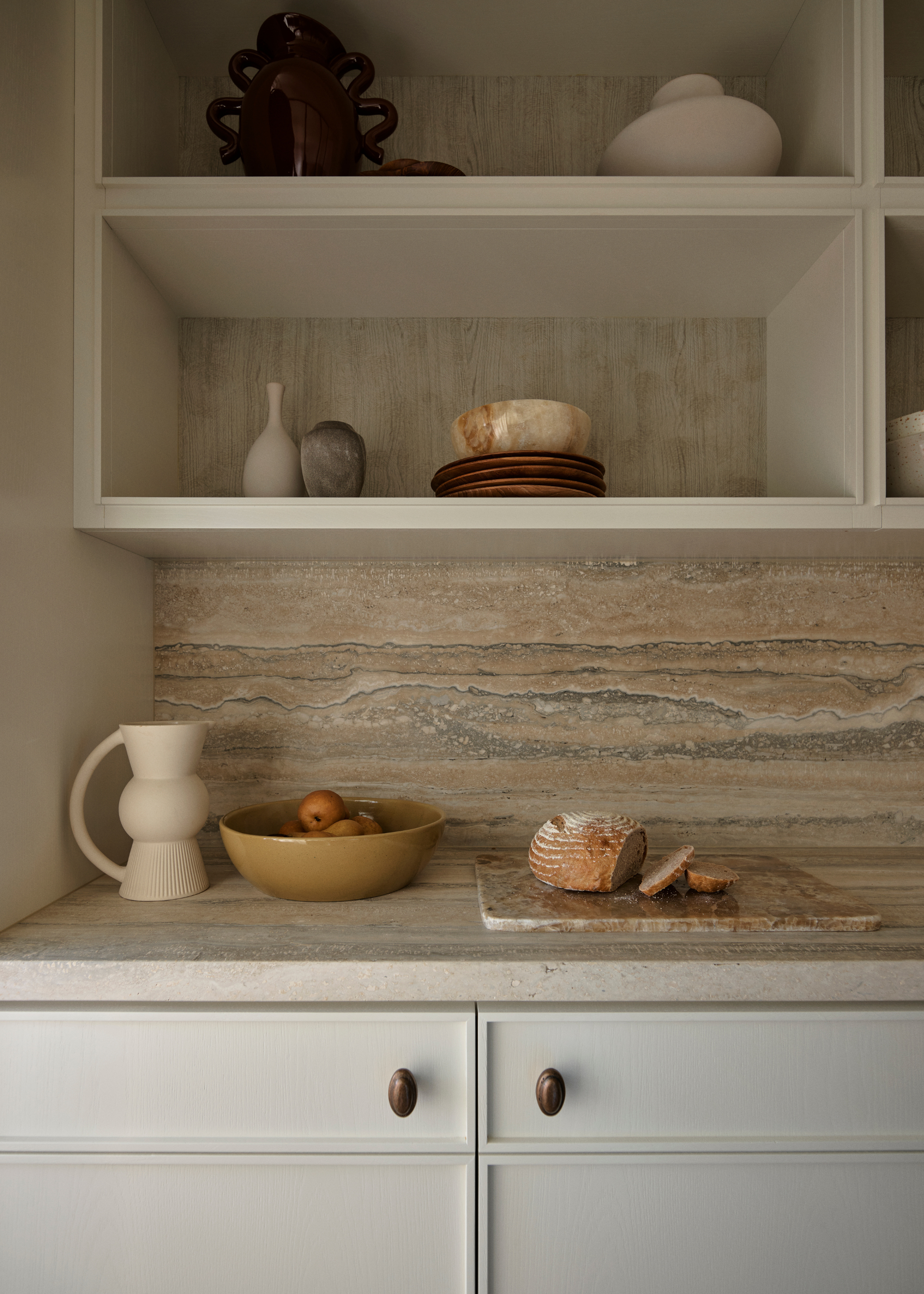 A kitchen with a stone backsplash and counter with a jug, a bowl of fruit and a cutting board with bread. An open shelving cabinet above with plates, bowls and vases
