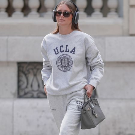 Diane Batoukina wears long, straight light brown hair styled with a middle part. Black Apple AirPods Max headphones and rectangular tortoiseshell Prada sunglasses frame the face. A heather grey UCLA sweatshirt is paired with matching Sporty  Rich sweatpants. A white ribbed crop top by Zara is worn underneath. Beige suede Miu Miu sneakers feature mismatched yellow and brown laces with white bows and tassel details. A grey leather Hermes Birkin bag is carried in hand, adorned with matte black AirPods Max headphones. A silver Rolex watch is worn on the left wrist. A green iced drink is held in the right hand. Full body shot, during a street style fashion photo session, on April 28, 2025 in Paris, France.