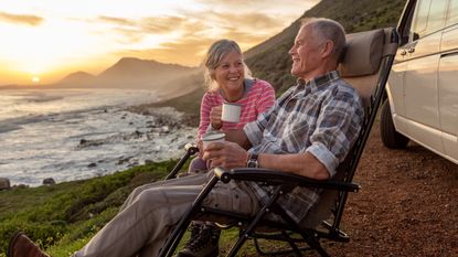 An older couple drink coffee while sitting on the waterfront outside their truck.