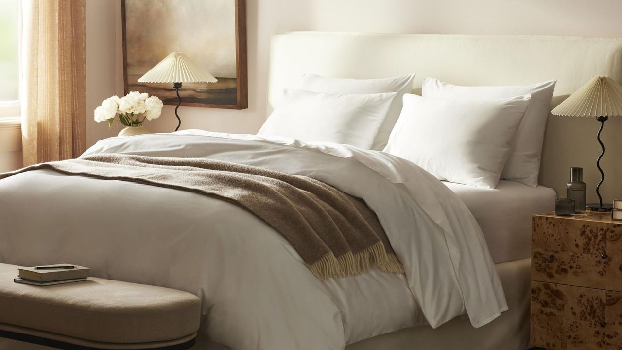 Brooklinen Luxury Sheet Set on a bed with a beige blanket; a nightstand either side, topped with a lamp and a vase of flowers; against a cream wall. 