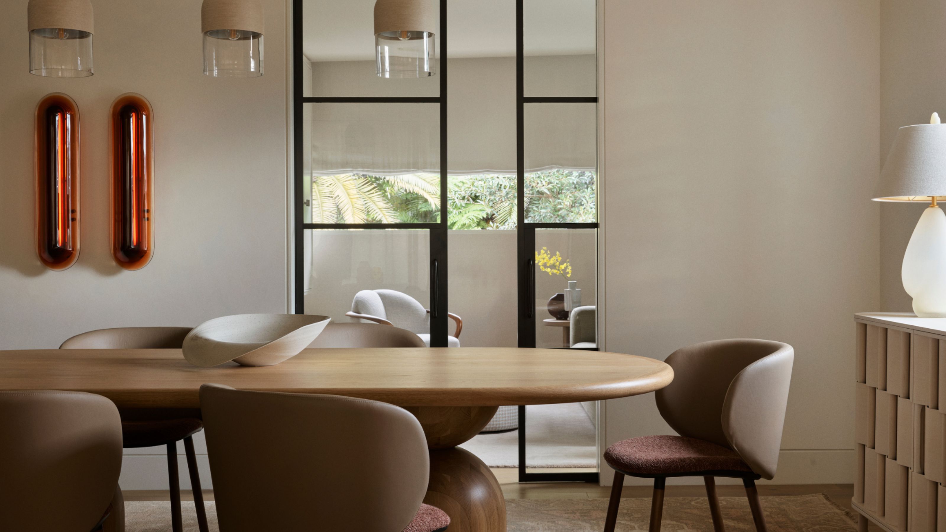 earth toned dining room with beige walls, dark brown chairs, timber table and console, white table lamp, orange wall art, and glass doors leading through to a living room with views of trees