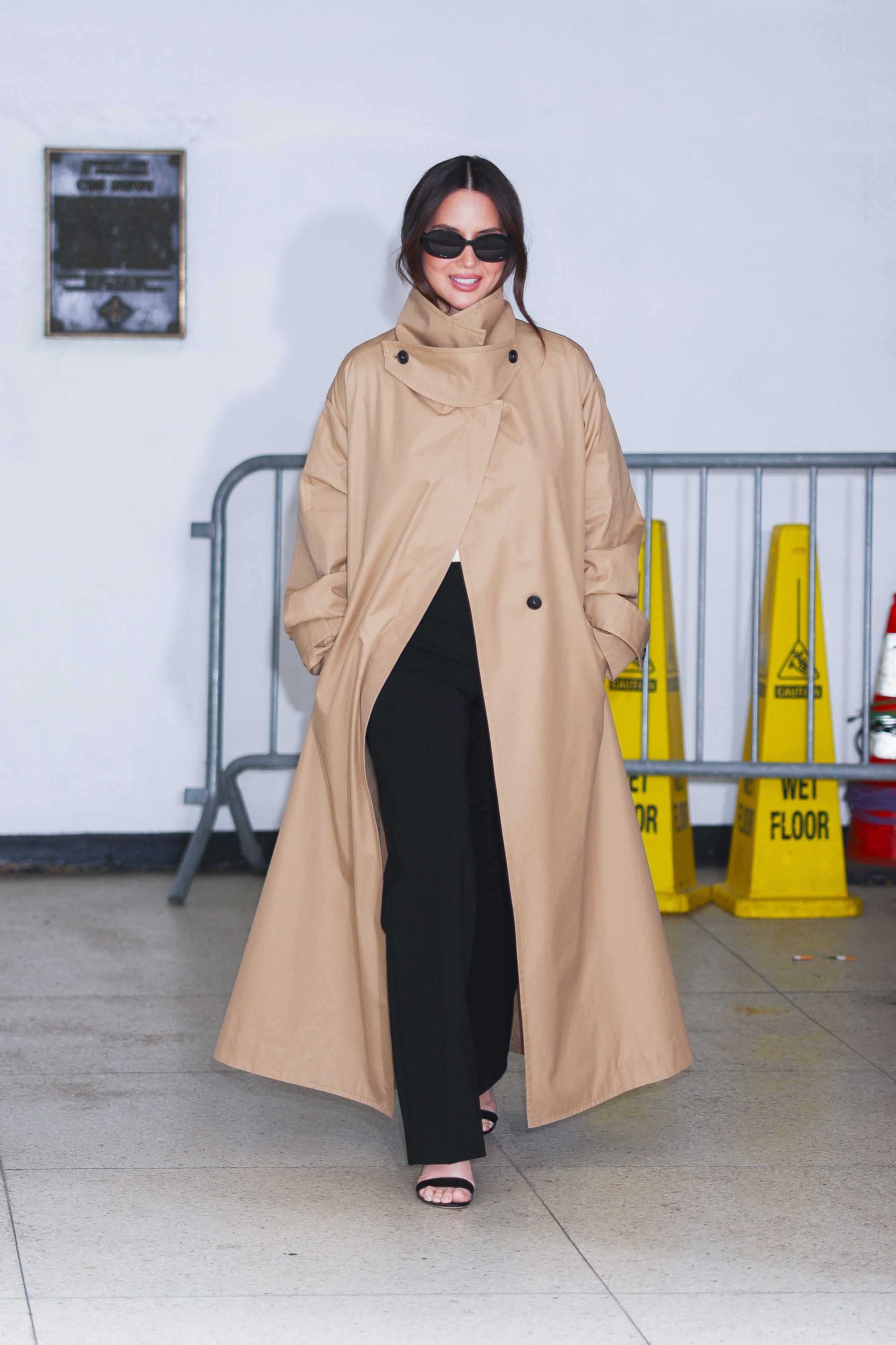 Olivia Munn wears a funnel neck trench coat with black trousers and black strappy sandals.