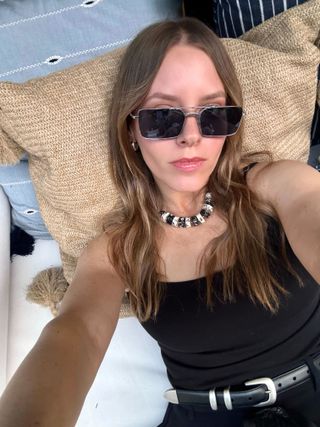 Nikki Chwatt wears Vogue Eyewear metal-framed sunglasses with a black and white necklace, black tank top, black belt, and black shorts.