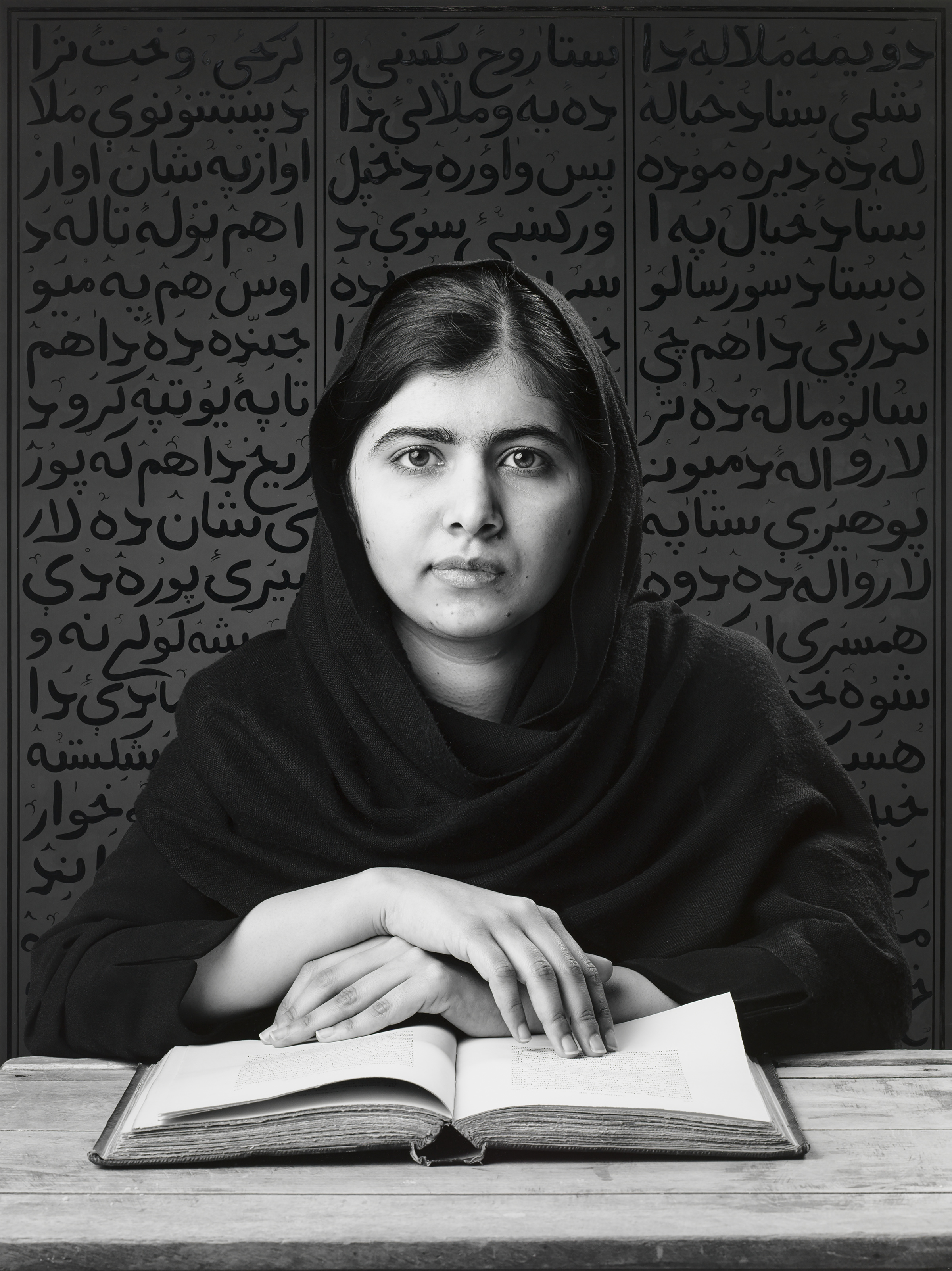 Malala Yousafzai's black-and-white portrait by Shirin Neshat, showing her seated at a desk with an open book, wearing a black hijab, with dense Pashtu calligraphy covering the background.